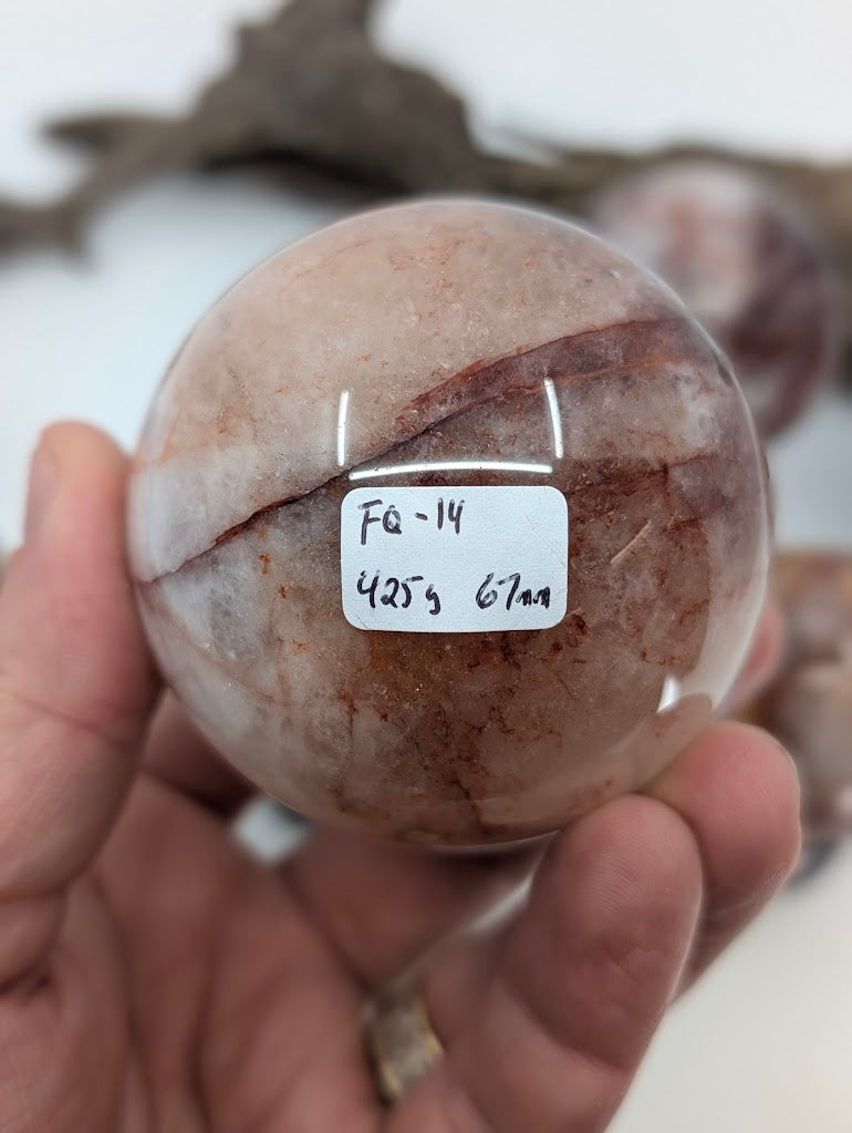 Collector hand scale verification photo of large Brazilian Fire Quartz sphere FQ-14, showing the tracking label marked 425 grams and 67mm.
