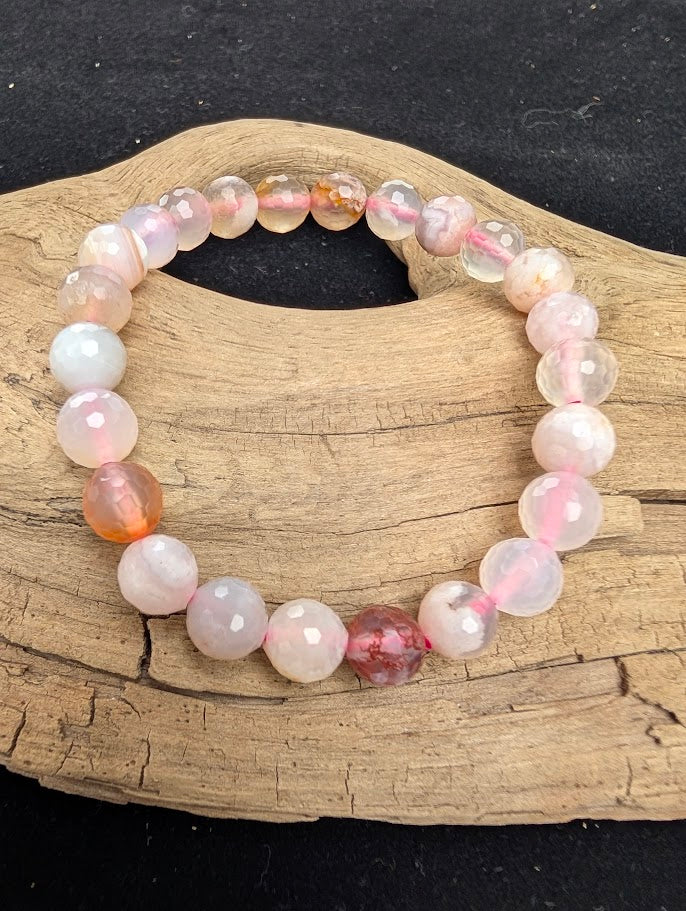 8mm faceted Flower Agate beaded stretch bracelet resting on a driftwood display, showing translucent stones with internal white and peach plume inclusions strung on pink cord.