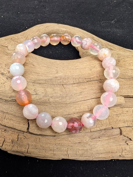8mm faceted Flower Agate beaded stretch bracelet resting on a driftwood display, showing translucent stones with internal white and peach plume inclusions strung on pink cord.