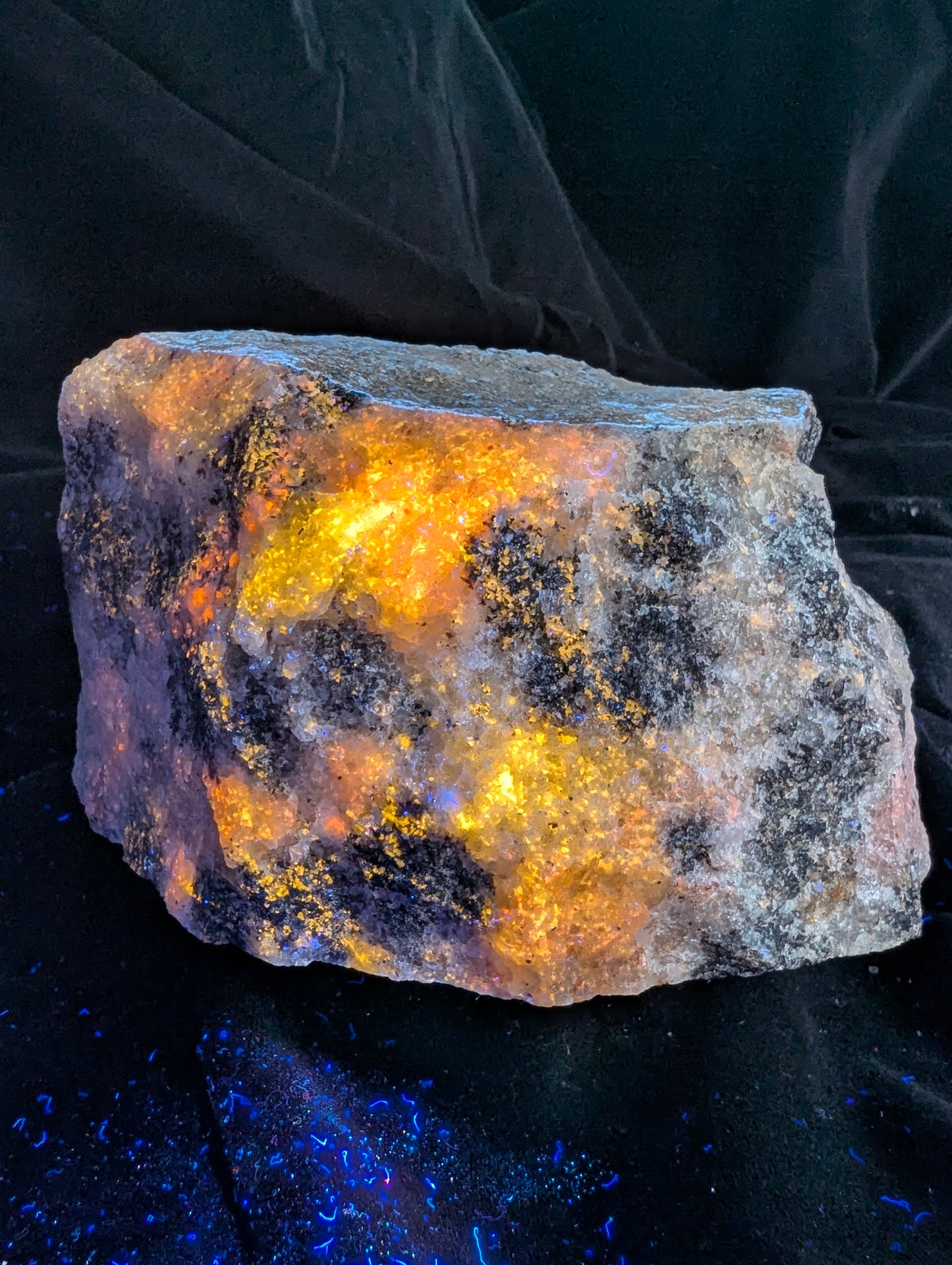 An angled view focusing on the bottom and front edges of the fluorescent Scapolite (Wernerite) specimen from Bancroft, Ontario, under UV light. The intense yellow and orange glow reflects slightly off the black velvet surface, displaying maximum fluorescent saturation and a rugged crystalline texture.