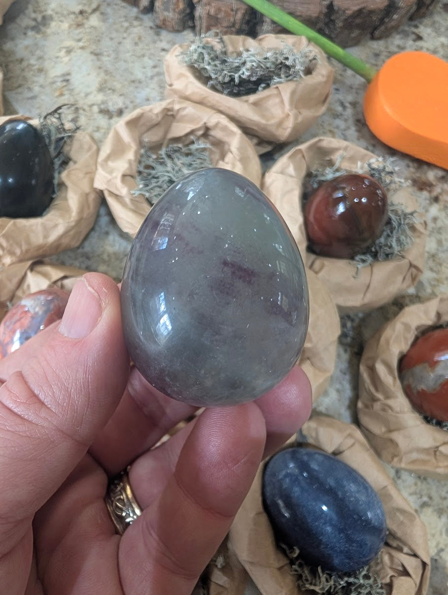 A 2-inch polished Fluorite egg held in a hand, displaying muted greyish-purple tones and smooth natural stone texture.