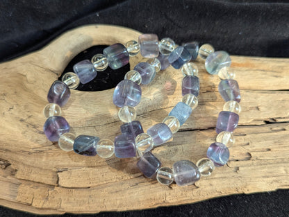 A 9 mm blue fluorite cube and 8 mm clear quartz round bead stretch bracelet positioned on a neutral presentation surface, demonstrating precise geometric lapidary cuts and high structural contrast.