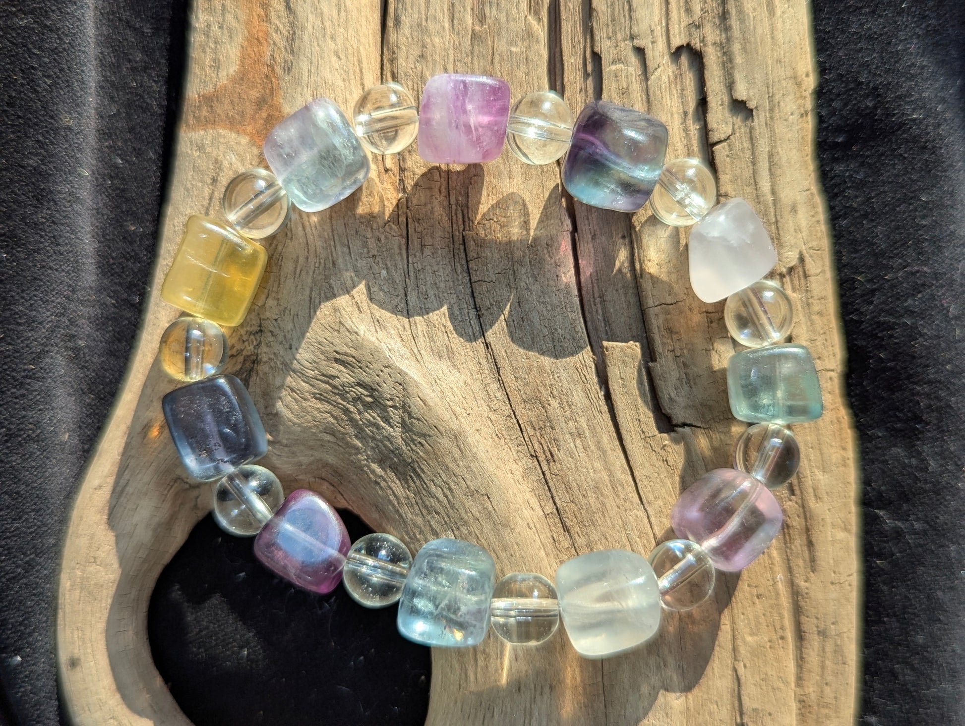 A 9 mm mixed-colour fluorite cube and 8 mm clear quartz round bead stretch bracelet resting on a neutral surface, displaying a vibrant spectrum of unaltered natural fluorite shades.