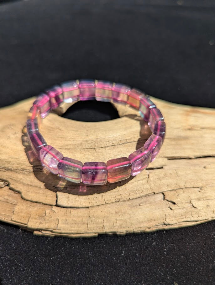 Polished Fluorite square segment stretch bracelet resting on a driftwood display, showing translucent purple and clear zones strung on a pink elastic cord.