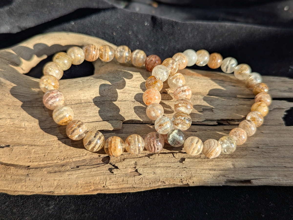 Two 8mm and 9mm Garden Quartz bracelets on a piece of driftwood, showing clear quartz beads with multi-dimensional peach and rust stacked phantom layers.