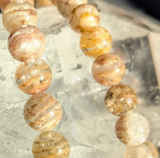 Macro close-up of Garden Quartz beads resting on a raw mineral, showing detailed horizontal terraced phantom layers in peach and sandy brown.