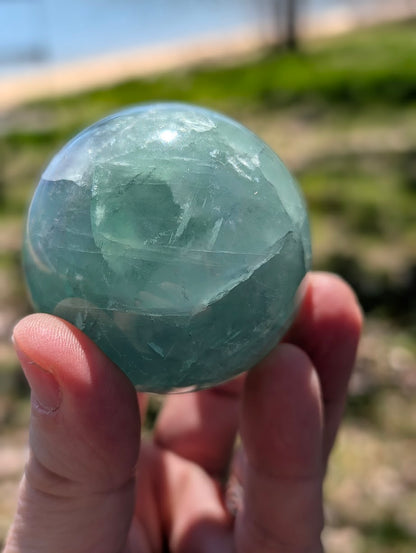 Hand holding a green fluorite sphere outdoors, showing its polished surface and deep internal mineral structure.