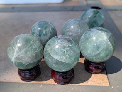 Hand holding a translucent green fluorite sphere in natural sunlight, highlighting its vibrant seafoam colouration.