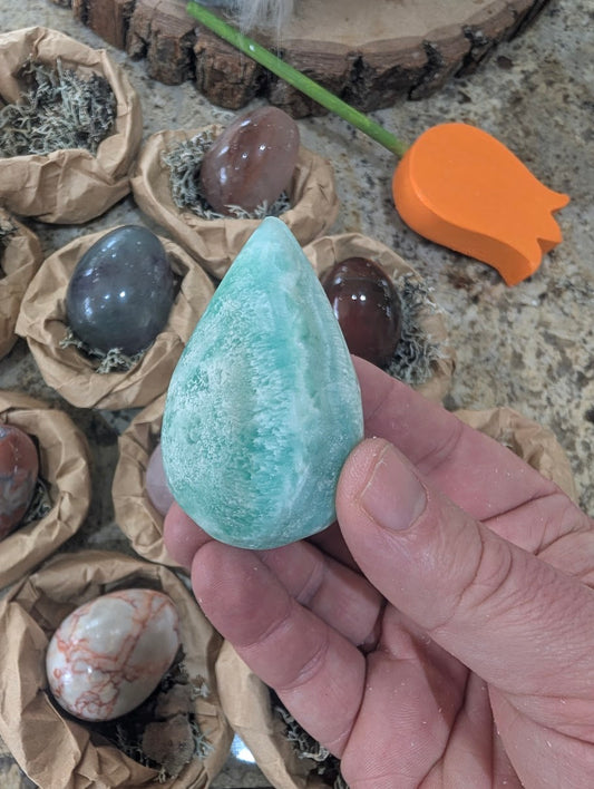 A 2-inch polished Hemimorphite egg held in a hand, displaying soft pale blue and teal tones mixed with natural white matrix textures.