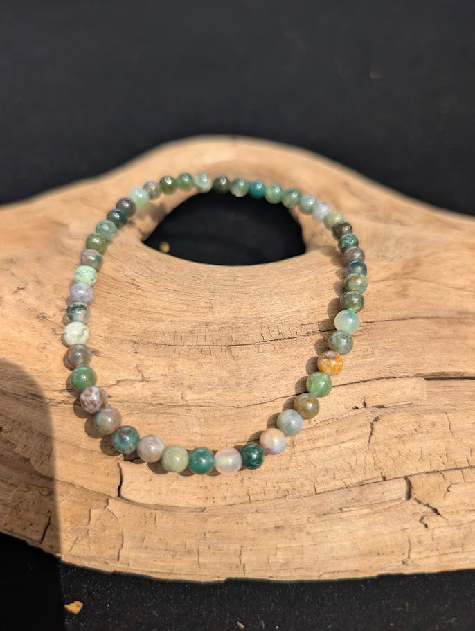 A 4mm Indian Agate beaded bracelet resting on natural driftwood, displaying earthy green, dusty purple, and tan colours.