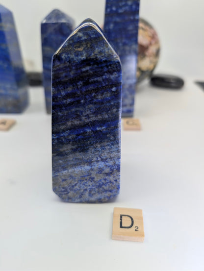 Polished Afghan Lapis Lazuli tower D (LT-04), standing 11cm tall, labeled with Scrabble tile D, featuring complex, wavy patterns of white calcite and prominent golden pyrite flecks against rich blue stone.