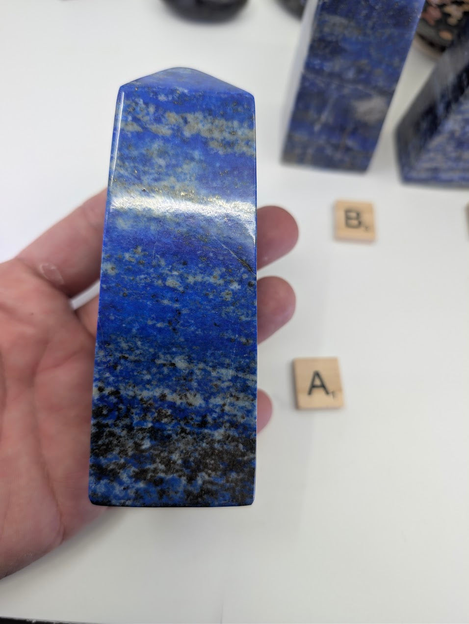 A close-up view of the polished Afghan Lapis Lazuli tower A (LT-01) held in a hand, displaying the natural contrast between the intense blue lazurite, metallic golden pyrite, and white calcite banding.