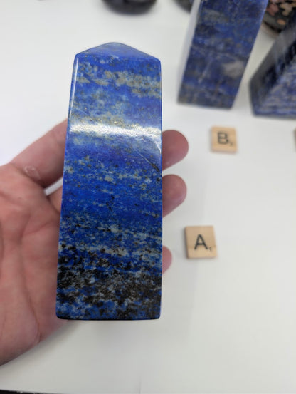 A close-up view of the polished Afghan Lapis Lazuli tower A (LT-01) held in a hand, displaying the natural contrast between the intense blue lazurite, metallic golden pyrite, and white calcite banding.