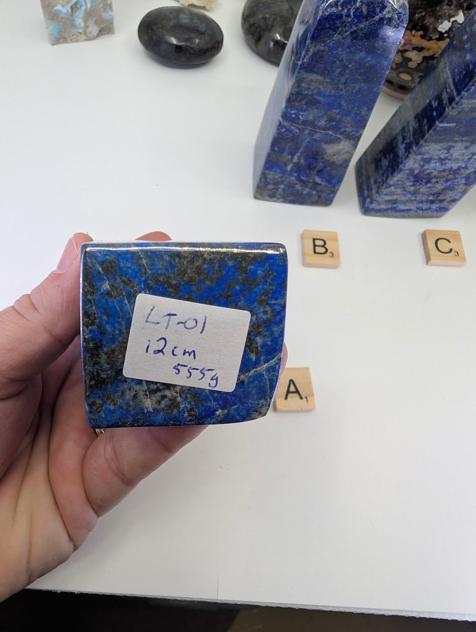 Hand-held view of the unpolished base of Lapis Lazuli tower LT-01 from Afghanistan, displaying the handwritten stock label "LT-01, 12cm, 555g" and the rich blue color of the stone matrix.