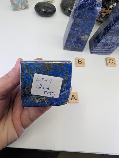 Hand-held view of the unpolished base of Lapis Lazuli tower LT-01 from Afghanistan, displaying the handwritten stock label "LT-01, 12cm, 555g" and the rich blue color of the stone matrix.