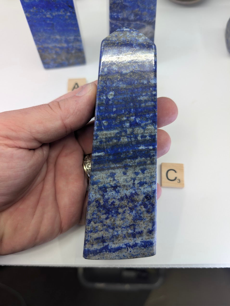 Afghan Lapis Lazuli tower C (LT-03) held in hand, showcasing the polished finish and the dynamic geological banding of white calcite and metallic golden pyrite within the ultramarine blue stone.