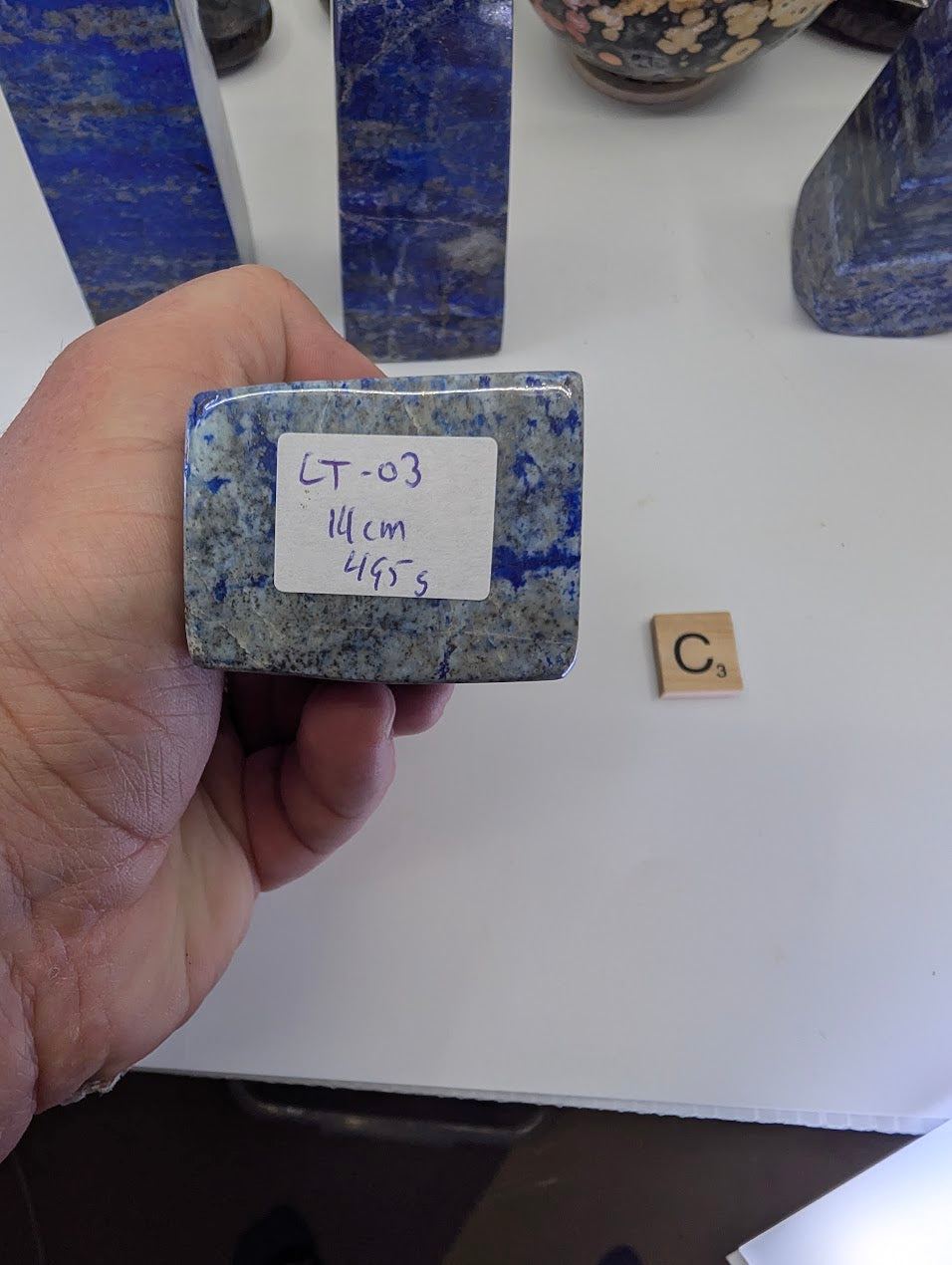 The flat, unpolished bottom surface of Afghan Lapis Lazuli tower LT-03, featuring a handwritten inventory tag labeled "LT-03, 14cm, 495g" against the granular texture of the stone matrix.