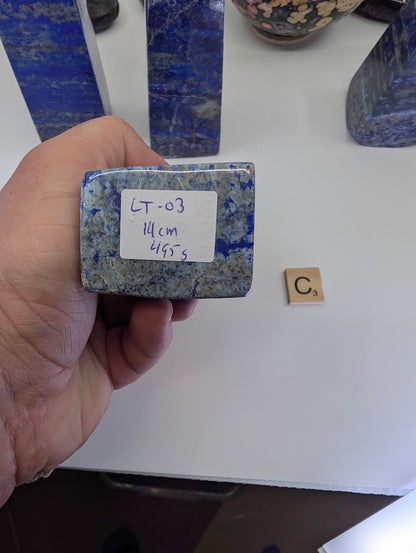 The flat, unpolished bottom surface of Afghan Lapis Lazuli tower LT-03, featuring a handwritten inventory tag labeled "LT-03, 14cm, 495g" against the granular texture of the stone matrix.
