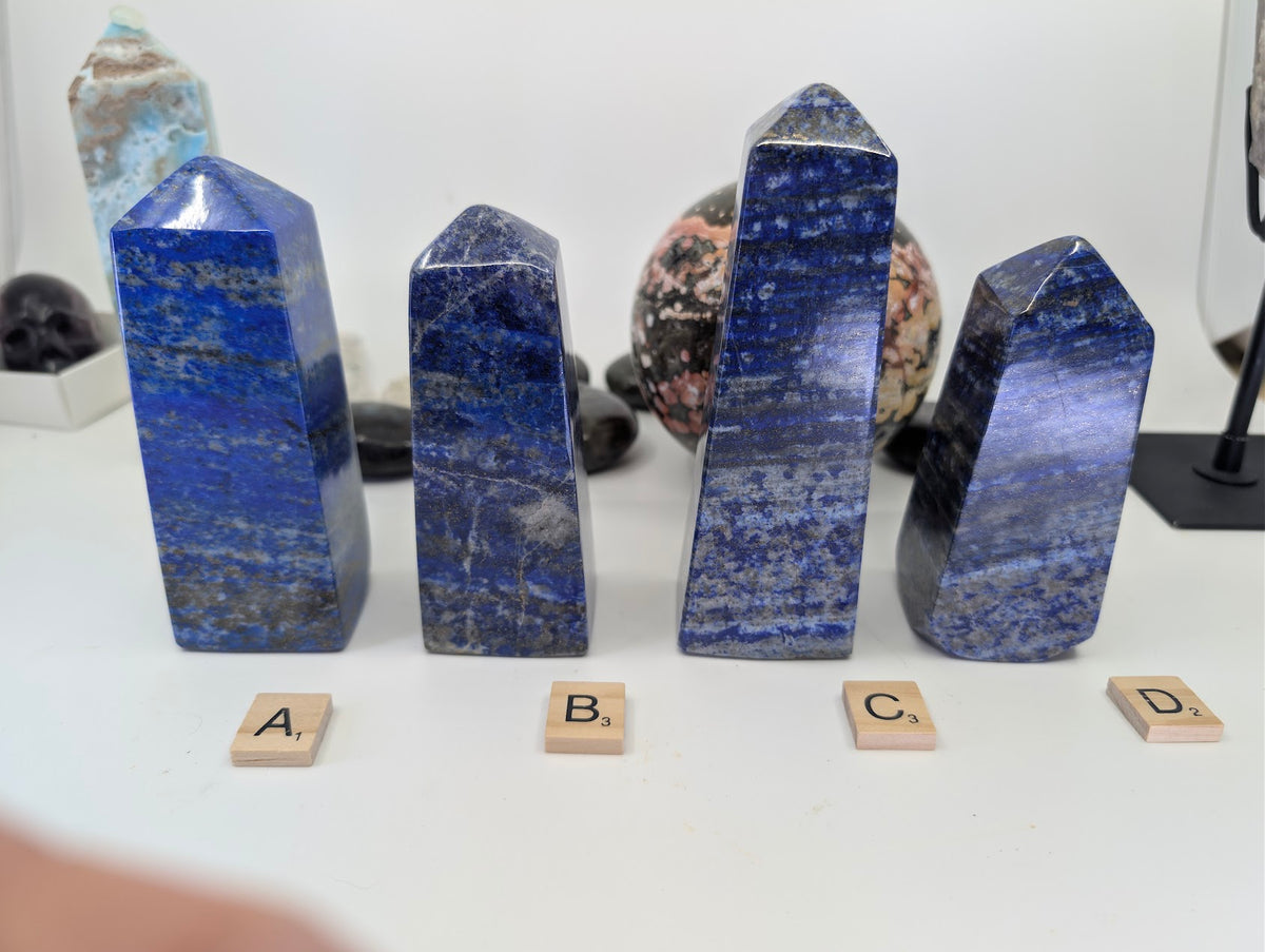 A group of four polished Lapis Lazuli towers from Afghanistan, labeled with Scrabble tiles A, B, C, and D, showing variations in rich blue color and calcite banding on a white background.