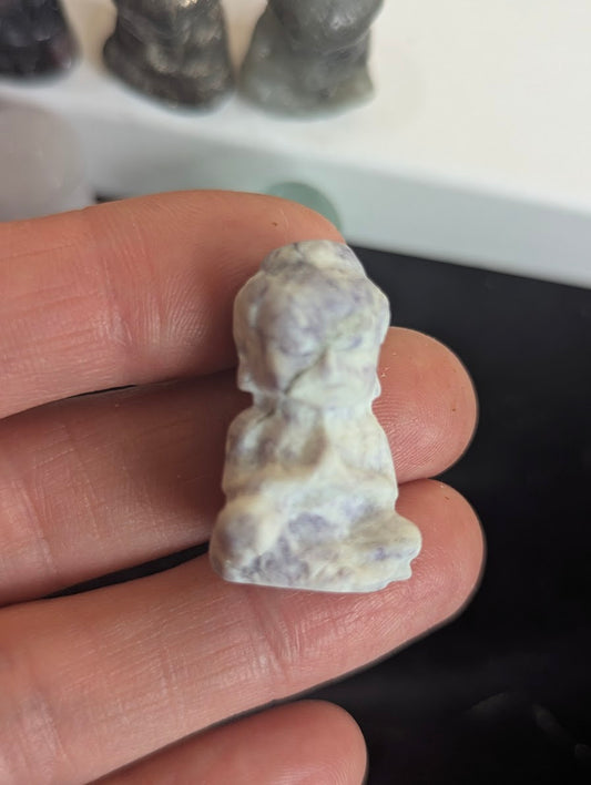 Handheld front view of a 3cm meditating monk totem carved from white matrix stone with purple lepidolite mica inclusions.