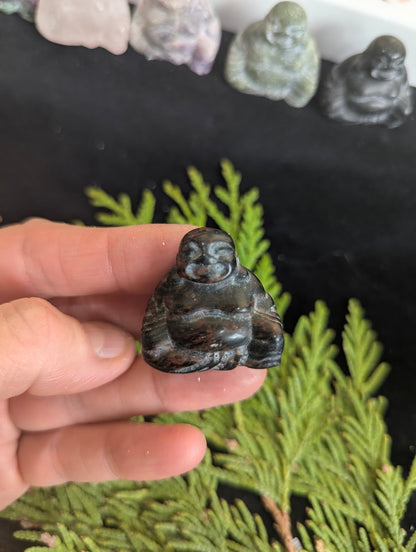 Close-up detail of a carved Mahogany Obsidian Hotei totem held against a green cedar branch background.