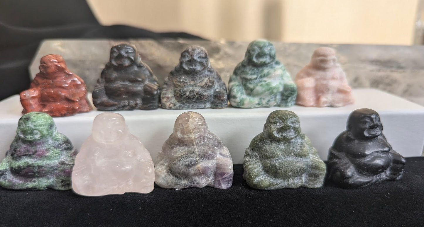 A row of ten mini Buddha totems carved from various natural minerals including Red Jasper, Amethyst, and Obsidian on a white and black display.