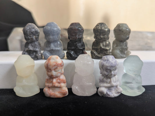 A group of ten mini meditating monk totems carved from various natural minerals including pyrite, fluorite, and agate on a display.