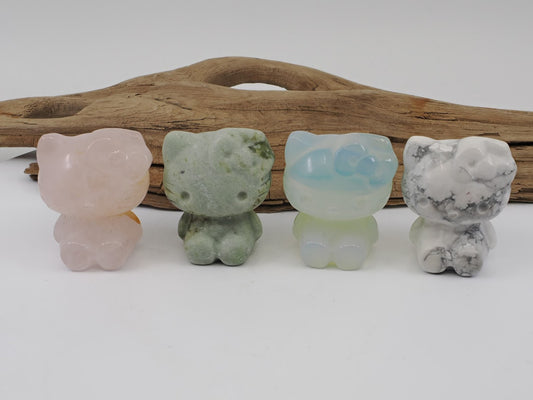 A collection of four Hello Kitty carvings in Rose Quartz, Green Aventurine, Opalite, and Howlite, standing 40mm tall in front of natural driftwood.