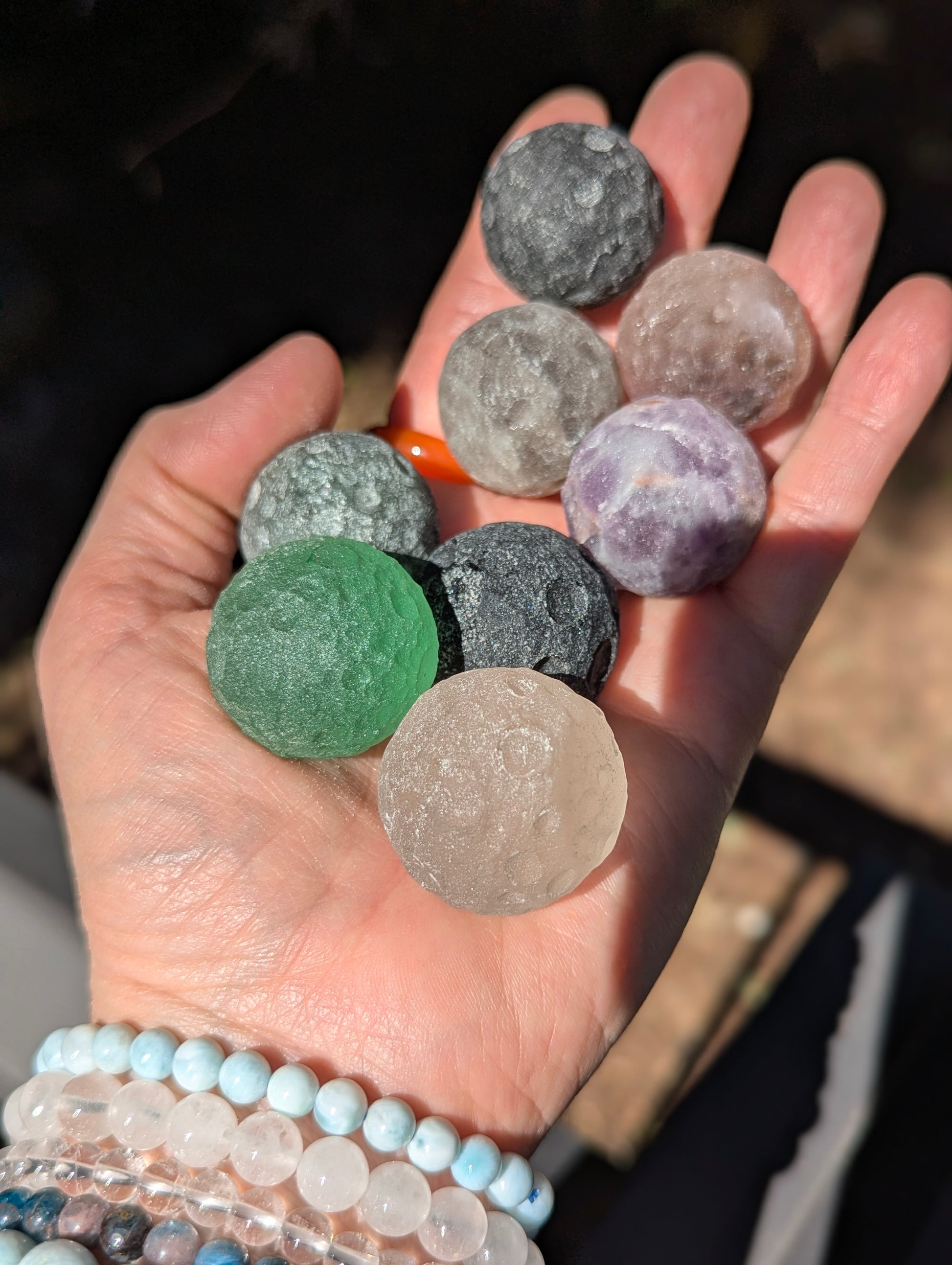 A hand holding a cluster of 30 mm (1.18 in) mini carved mineral moon spheres, including Green Aventurine, Black Obsidian, Amethyst, and Rose Quartz.