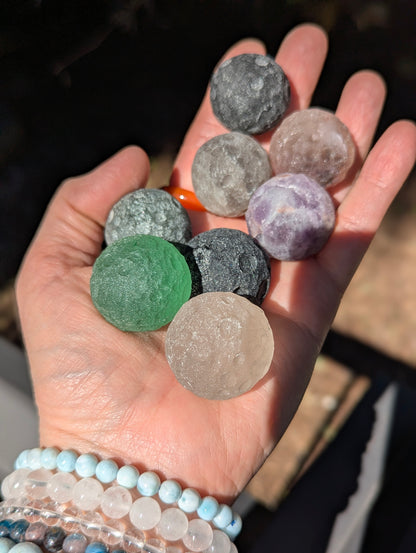 A hand holding a cluster of 30 mm (1.18 in) mini carved mineral moon spheres, including Green Aventurine, Black Obsidian, Amethyst, and Rose Quartz.