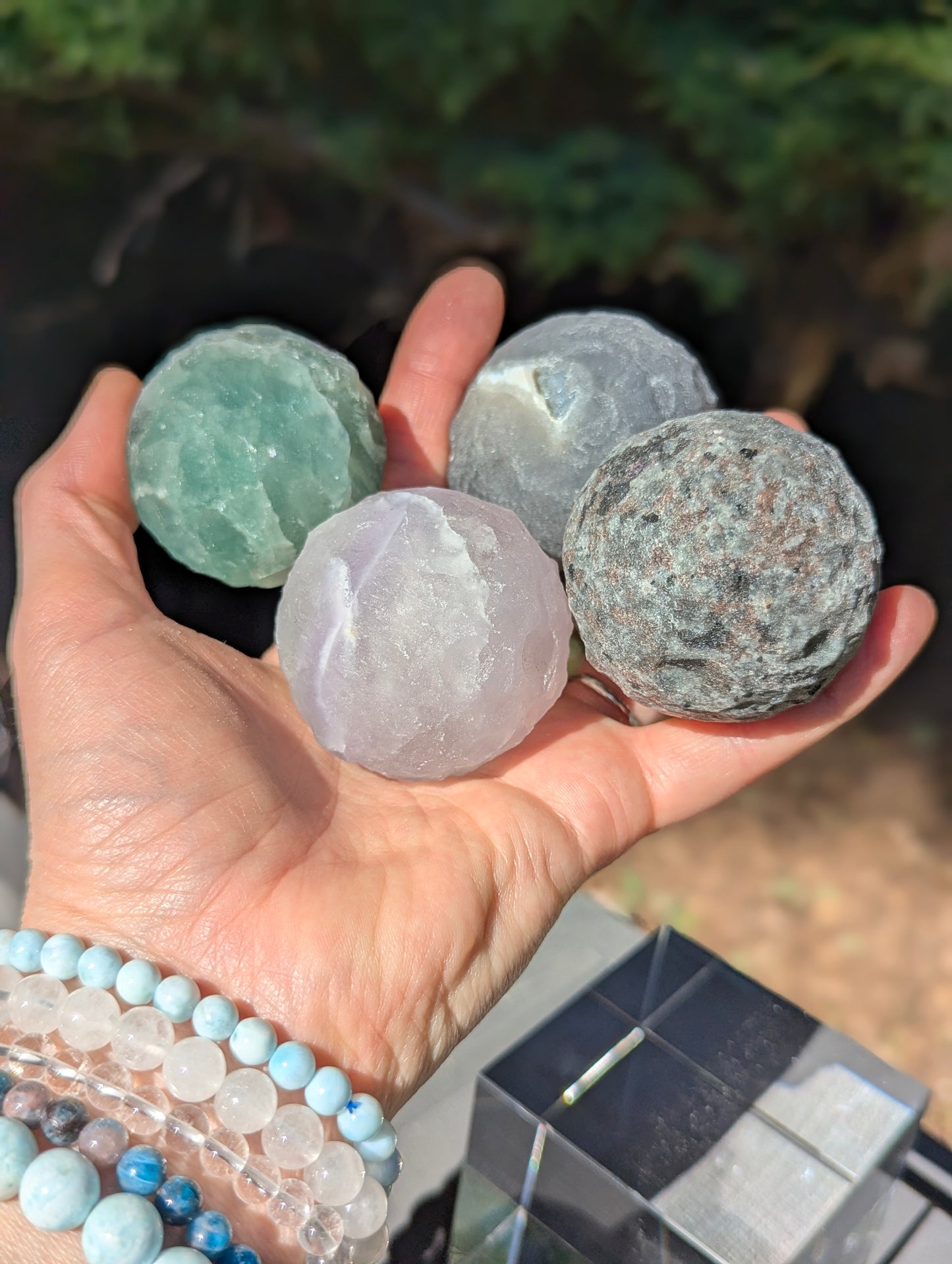 A hand holding four 50 mm (1.97 in) carved mineral moon spheres, including Green Aventurine, Silk Fluorite, Larvikite, and Yooperlite, in natural light.