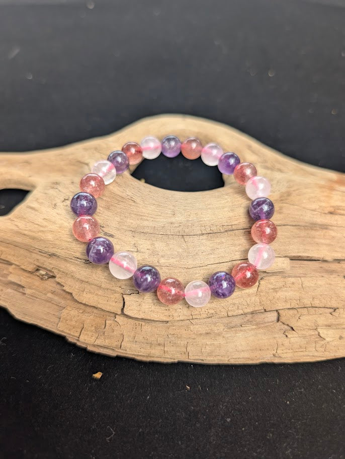 An 8mm mixed quartz beaded bracelet resting on natural driftwood, displaying purple Amethyst, soft pink Rose Quartz, and speckled Strawberry Quartz.