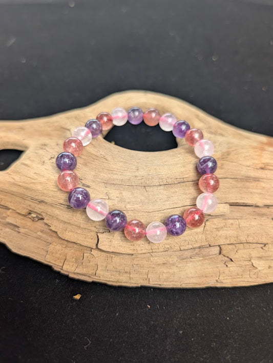 An 8mm mixed quartz beaded bracelet resting on natural driftwood, displaying purple Amethyst, soft pink Rose Quartz, and speckled Strawberry Quartz.