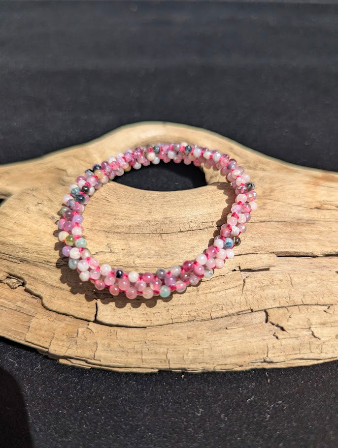 Hand-woven mixed Tourmaline beaded bracelet resting on a driftwood display, showing pink, green, and black micro-beads intricately woven on a pink cord.