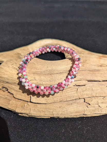 Hand-woven mixed Tourmaline beaded bracelet resting on a driftwood display, showing pink, green, and black micro-beads intricately woven on a pink cord.