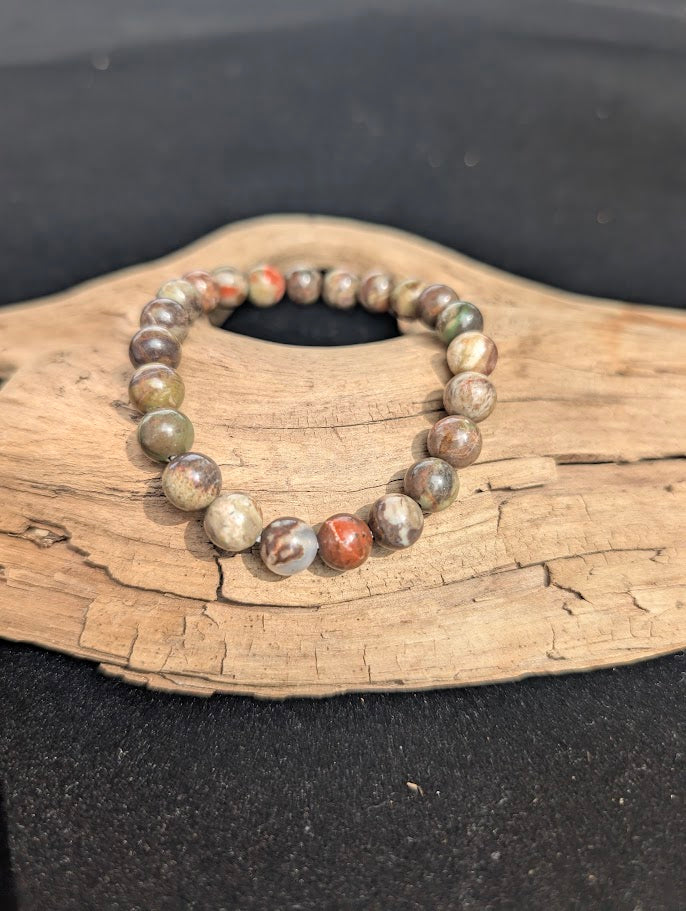 An 8mm Money Agate beaded bracelet resting on natural driftwood, displaying earthy red, green, and grey chalcedony colours.