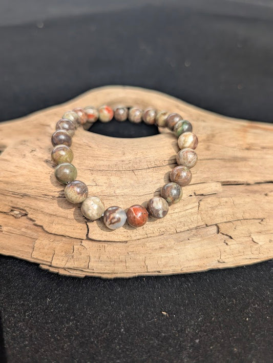 An 8mm Money Agate beaded bracelet resting on natural driftwood, displaying earthy red, green, and grey chalcedony colours.