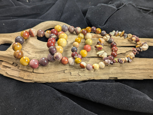 A complete collection of authentic Mookaite stretch bracelets from Western Australia in chip, 8mm, 10mm, and 12mm bead sizes, resting on natural driftwood. The group shot showcases an earthy, un-stabilized radiolarite matrix featuring high-contrast burgundy, mustard, and cream tones.
