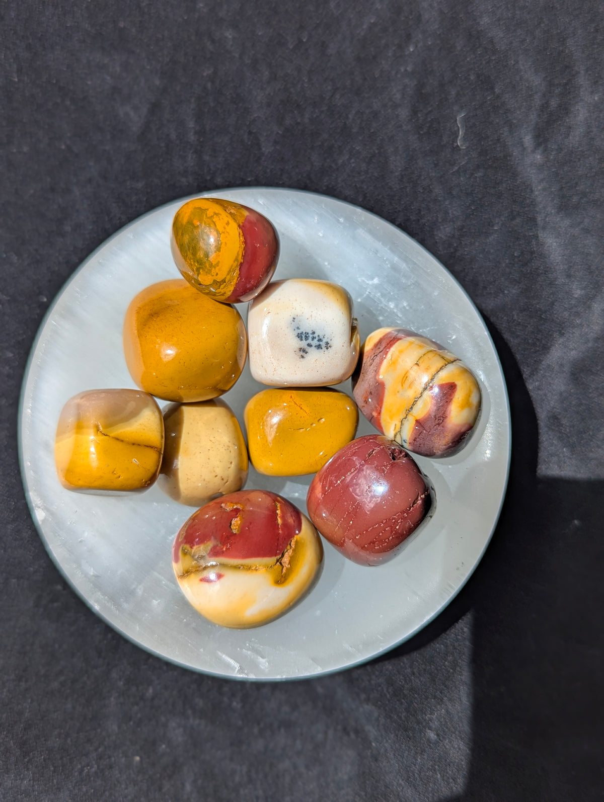 Nine polished Mookaite tumbled stones displaying yellow, red, and cream banding resting on a white display plate.