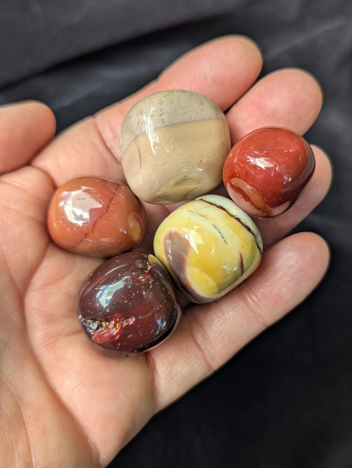Close-up of five high-gloss Mookaite tumbled stones held in a palm, showing natural surface vugs and vibrant colour banding.