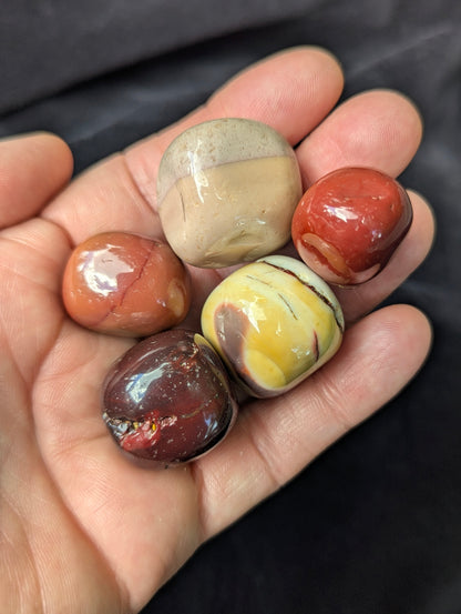Close-up of five high-gloss Mookaite tumbled stones held in a palm, showing natural surface vugs and vibrant colour banding.