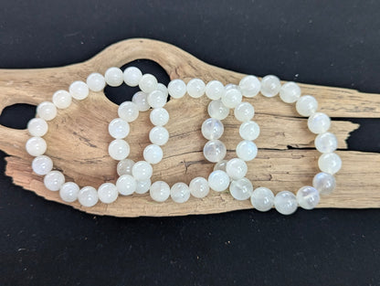 Three sizes of cloudy white Sri Lankan Moonstone bracelets on driftwood, showcasing 10mm, 11mm, and 12mm variants.