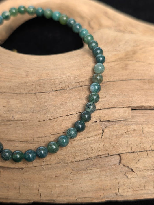 Macro view of polished 4mm Moss Agate spherical beads on driftwood, highlighting the natural green chlorite dendrites suspended in clear quartz.