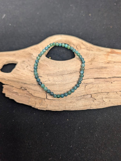 A 4mm Moss Agate beaded bracelet resting on natural driftwood, displaying translucent chalcedony with deep green botanical inclusions.