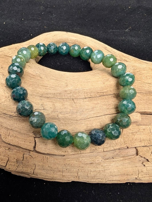 8mm faceted Moss Agate beaded stretch bracelet resting on a driftwood display, showing translucent chalcedony with dark forest green dendritic inclusions.