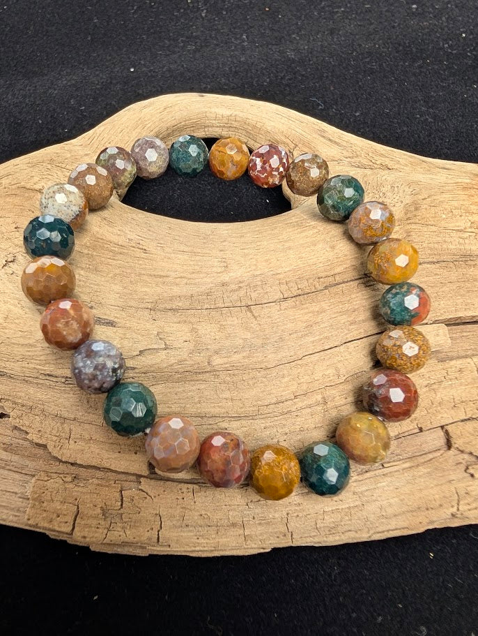 8mm faceted Ocean Jasper beaded stretch bracelet resting on a driftwood display, showing vibrant teal, mustard, and earthy red orbicular patterns.