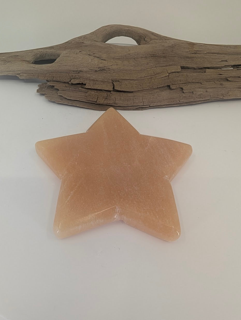 Polished Orange Calcite star displayed on a white surface in front of natural driftwood, highlighting the stone's symmetrical points and warm amber hue.