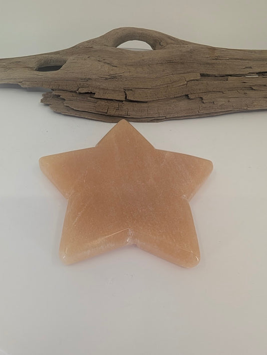 Polished Orange Calcite star displayed on a white surface in front of natural driftwood, highlighting the stone's symmetrical points and warm amber hue.