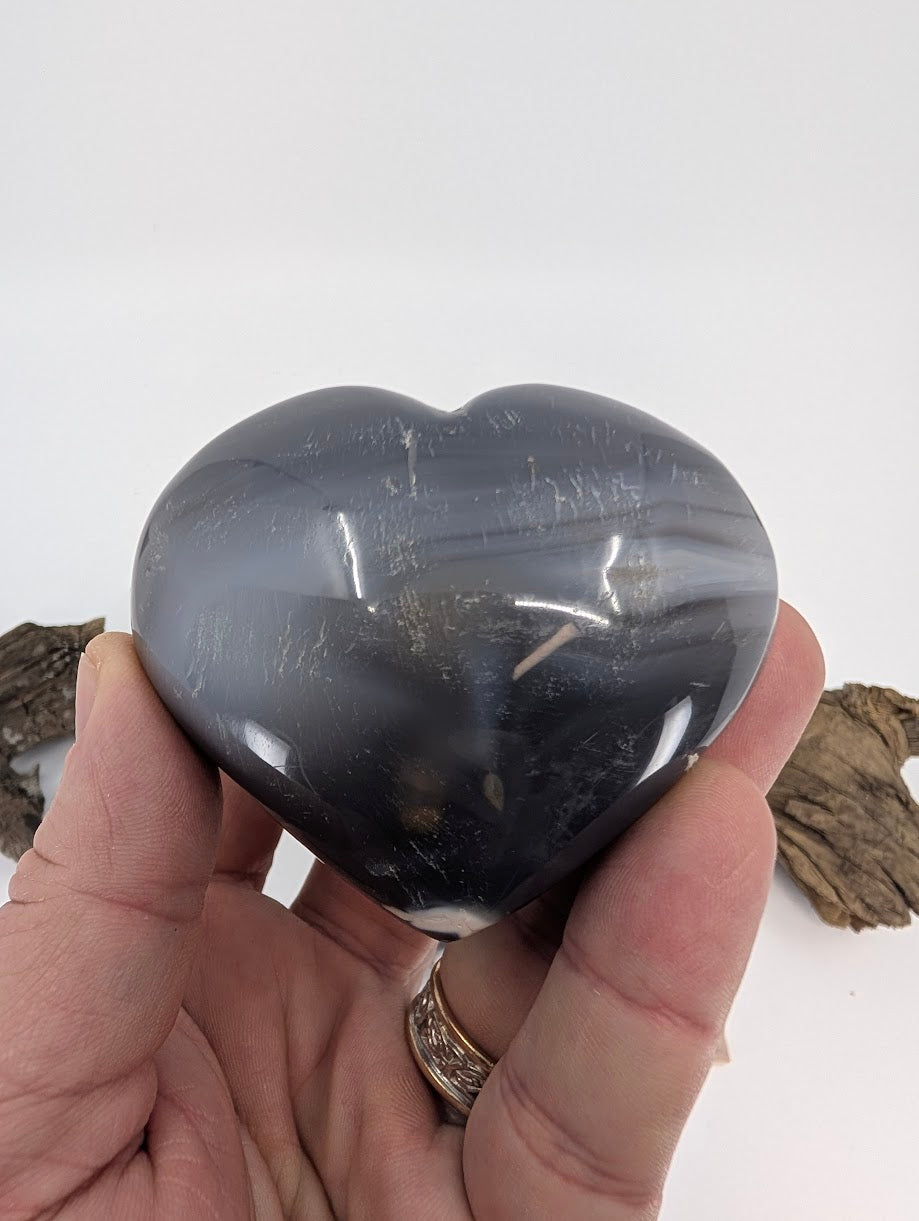Hand holding Specimen C, a dark moody Orca Agate heart.