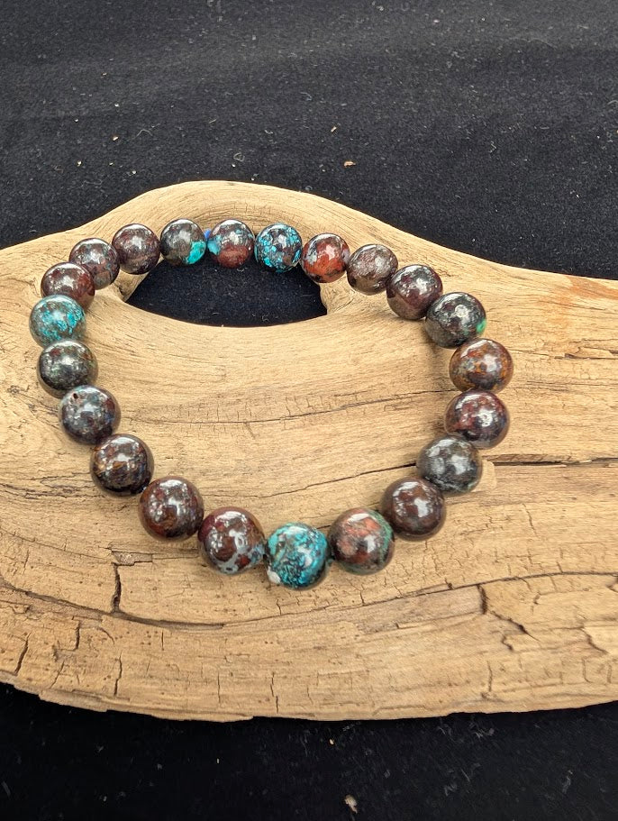 8mm polished Phoenix Stone beaded stretch bracelet resting on a driftwood display, showing a dark copper matrix with teal and green inclusions.